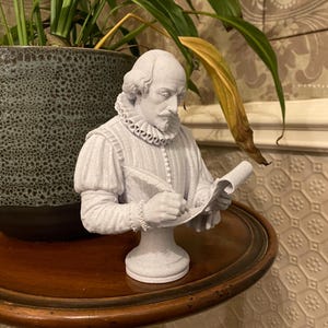 May include: A light grey bust of William Shakespeare depicted in a historical doublet with a ruffled collar. The figure is shown in profile, holding a quill pen and writing on a scroll. The sculpture features a textured, matte finish and rests on a pedestal base.