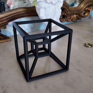 May include: A matte black geometric sculpture featuring a tesseract design, which consists of a smaller cube nested within a larger cube, connected by straight rods at each corner to form a three-dimensional wireframe structure of a four-dimensional hypercube.