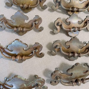 May include: Eight brass drawer pulls arranged in two columns. Each pull features a decorative backplate with curved edges and a central swinging bail handle. The metal has a tarnished, aged finish with visible patina and subtle green oxidation.