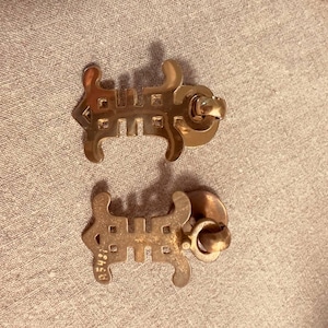 May include: Two gold-toned metal drawer pulls featuring a stylized, symmetrical geometric design with a central vertical bar and outward-curving ends. Each pull includes a circular mounting base and a small, attached ring handle for gripping.