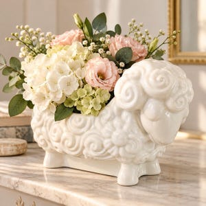 May include: A white ceramic sheep-shaped planter featuring a glossy finish and embossed swirl patterns on its body. The planter is filled with a floral arrangement of light pink roses, white hydrangeas, green foliage, and small white filler flowers.