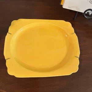 May include: A bright yellow square plate with rounded corners and a smooth, glossy finish. The plate features a wide, flat rim and a central circular depression. It is held in place by four thin, silver-toned metal clips attached to the edges.