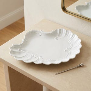May include: A white ceramic serving platter with an irregular, scalloped edge and embossed scrollwork details along the rim. The platter features a smooth, glossy finish and is accompanied by a slender, silver-toned metal serving pick.