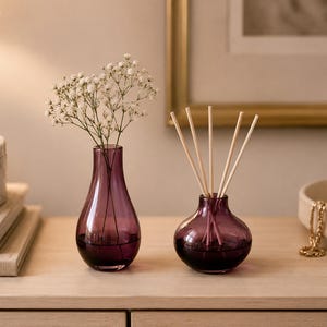 May include: Two purple glass vessels with a smooth, rounded shape. The taller vessel holds a small bouquet of white baby's breath flowers. The shorter, wider vessel contains five light-colored wooden reed diffuser sticks protruding from the top.