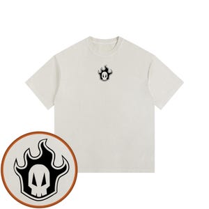 Puede incluir: Una camiseta de manga corta y cuello redondo en color blanco roto con un pequeño gráfico centrado en el pecho. El gráfico muestra una calavera blanca estilizada dentro de una forma de llama negra, con bordes blancos y negros.