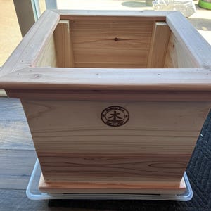 Könnte beinhalten: Ein quadratischer Pflanzkasten aus Holz mit hellbraunem Finish. Der Pflanzkasten hat einen dekorativen Rand und ein kreisförmiges Logo auf der Vorderseite. Er steht auf einer transparenten Kunststoffschale. Der Pflanzkasten ist für den Außen- oder Innenbereich konzipiert.