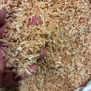 Könnte beinhalten: Nahaufnahme eines Haufens hellbrauner Holzspäne. Die Späne sind fein zerkleinert und wirken trocken. Eine Hand hält einen Teil der Späne und zeigt die Textur und Details des Materials. Der Hintergrund ist mit weiteren Holzspänen gefüllt.