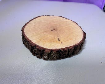 May include: A round wooden slice with a light brown interior and a dark brown, textured bark edge. The wood slice is likely a coaster or decorative item, showcasing natural wood grain.