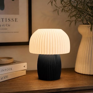May include: A modern table lamp with a black base and a cream-colored, ribbed lampshade. The lamp is on a wooden surface, with a framed print and books in the background. The lamp's design is minimalist and stylish.