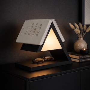 May include: A black triangular table lamp with a book-shaped shade. The book reads "GOOD VIBES, GOOD LIFE" and rests above a warm light. A pair of glasses sits on the base of the lamp.