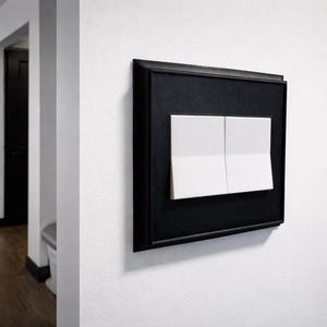 May include: A modern light switch with two white rectangular buttons set in a black frame, mounted on a white wall. The switch is part of a contemporary home interior, with a glimpse of a hallway and kitchen.
