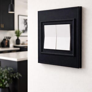 May include: A black light switch with two white switches, set in a black frame, mounted on a white wall. The switch is part of a modern kitchen design, with a black cabinet and a pendant light visible in the background.