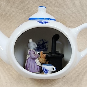 May include: A white ceramic teapot with a hollowed-out front displaying a miniature scene inside. The scene features a grey mouse figure in a purple dress standing next to a black stove, a small basket, and a tiny blue and white floral teacup.