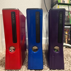 Peut inclure: Trois consoles Xbox 360 rouges, bleues et violettes. Chaque console a une fente noire pour les disques et le logo Xbox 360. Les consoles sont sur un tapis beige.