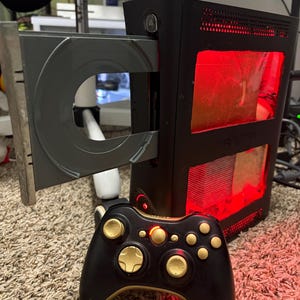 May include: Black Xbox 360 console with an open disc tray, illuminated by red internal lighting. A black and gold controller is in front of the console, featuring gold accents on the buttons and thumbsticks. The console is on a carpeted surface.