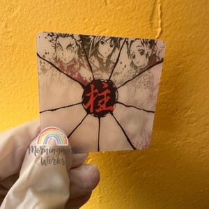 May include: A square, semi-translucent sticker featuring three anime-style character portraits arranged in the upper section. A central black circle contains the red Japanese kanji character for Hashira, with black lines radiating outward.