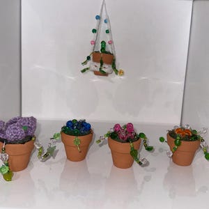May include: A collection of miniature terracotta pots, each containing a different handcrafted plant. The plants are made of various materials, including crochet and beads, and feature colors like purple, blue, pink, and orange. One pot is suspended from above.
