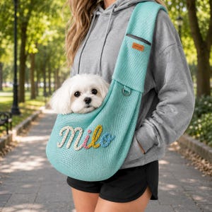 Puede incluir: Un transport&iacute;n de malla azul claro para mascotas con el nombre Milo bordado en cursiva con hilo blanco, rosa, amarillo y azul. El transport&iacute;n tiene una peque&ntilde;a etiqueta rectangular de cuero marr&oacute;n cerca de la correa y una anilla en D.