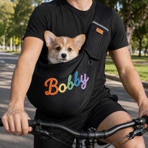 Puede incluir: Un transport&iacute;n de malla negra para mascotas con el nombre Bobby bordado en letras de hilo de colores. El transport&iacute;n tiene una correa ancha para el hombro con una peque&ntilde;a etiqueta rectangular marr&oacute;n y una anilla en D plateada, con un cachorro.