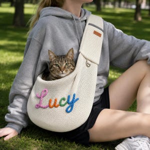 Puede incluir: Un transport&iacute;n de malla color crema para mascotas con el nombre Lucy bordado en letras de lana de colores. El transport&iacute;n tiene una correa ancha para el hombro con una peque&ntilde;a etiqueta rectangular marr&oacute;n y una anilla en D met&aacute;lica para un gato atigrado.