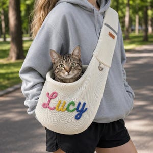 Puede incluir: Un transport&iacute;n de malla color crema para mascotas con el nombre Lucy bordado en letras de hilo de colores en la parte delantera. El transport&iacute;n tiene una correa ancha para el hombro con una peque&ntilde;a etiqueta rectangular marr&oacute;n y una anilla en D.