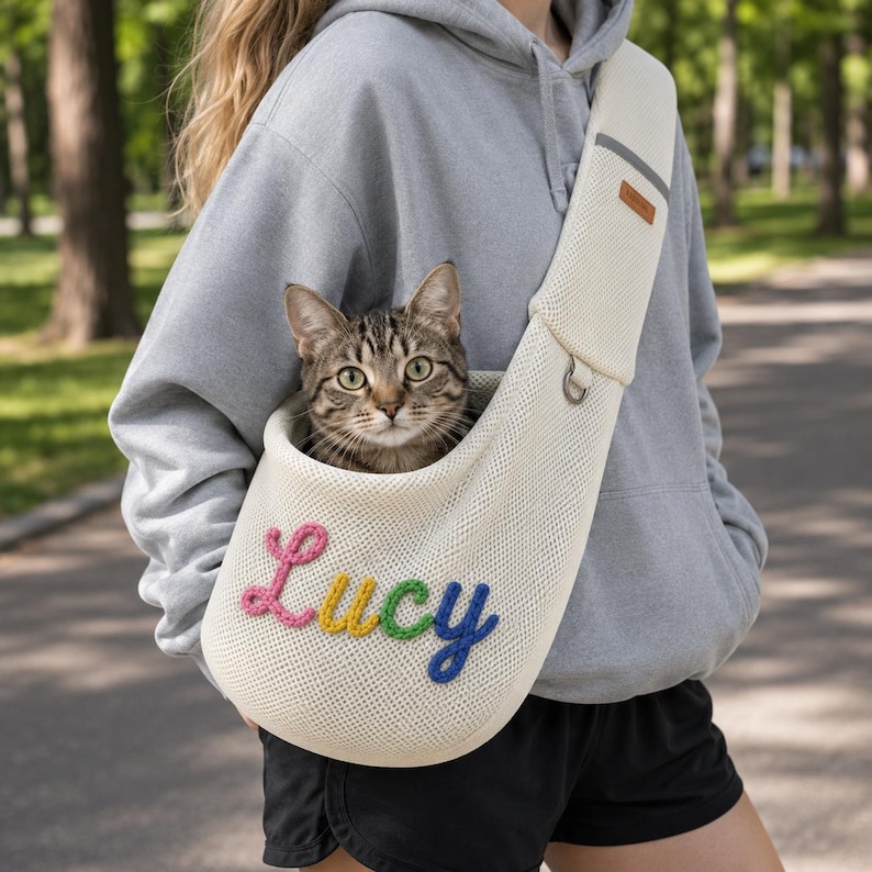 Puede incluir: Un transport&iacute;n de malla color crema para mascotas con el nombre Lucy bordado en letras de hilo de colores en la parte delantera. El transport&iacute;n tiene una correa ancha para el hombro con una peque&ntilde;a etiqueta rectangular marr&oacute;n y una anilla en D.