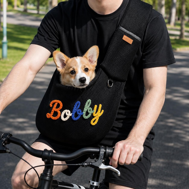 Puede incluir: Un transport&iacute;n de malla negra para mascotas con el nombre Bobby bordado en letras de hilo de colores. El transport&iacute;n tiene una correa ancha para el hombro con una peque&ntilde;a etiqueta rectangular marr&oacute;n y una anilla en D, con un cachorro Corgi.