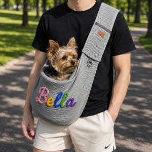 Puede incluir: Un transport&iacute;n de malla gris para mascotas con una correa ancha para el hombro y una peque&ntilde;a etiqueta rectangular marr&oacute;n. El nombre Bella aparece en la parte frontal en letras cursivas bordadas, coloridas y texturizadas. Un perro peque&ntilde;o.