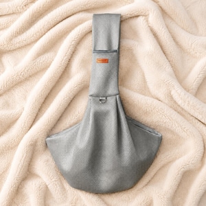 Puede incluir: Un portamascotas de malla gris con una correa ancha para el hombro, una peque&ntilde;a etiqueta rectangular naranja con la palabra FASHION impresa en negro y un anillo en D de metal situado en el centro del cuerpo estructurado en forma de bolsa.
