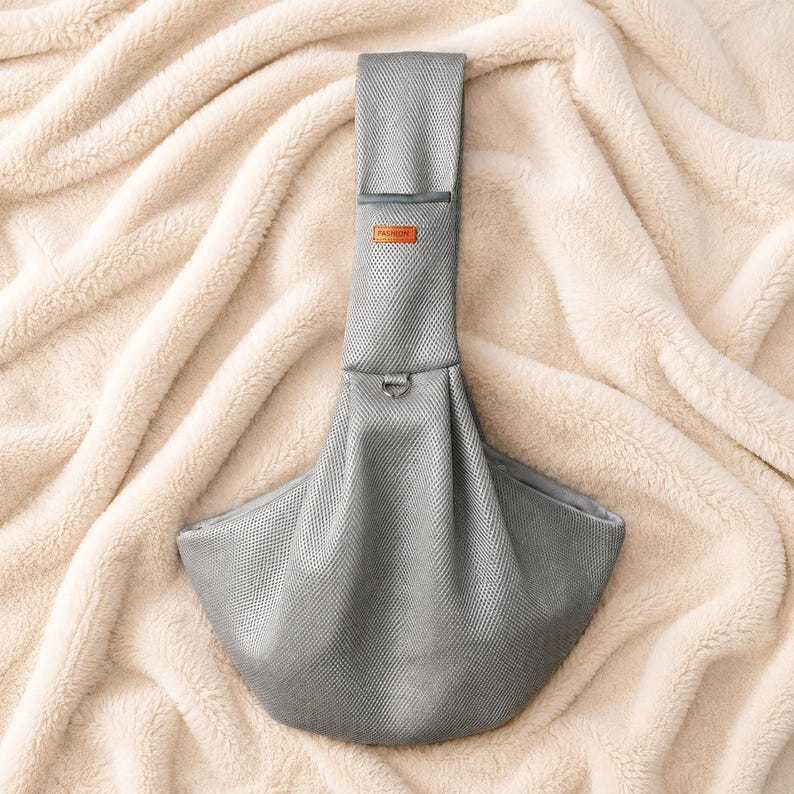 Puede incluir: Un portamascotas de malla gris con una correa ancha para el hombro, una peque&ntilde;a etiqueta rectangular naranja con la palabra FASHION impresa en negro y un anillo en D de metal situado en el centro del cuerpo estructurado en forma de bolsa.