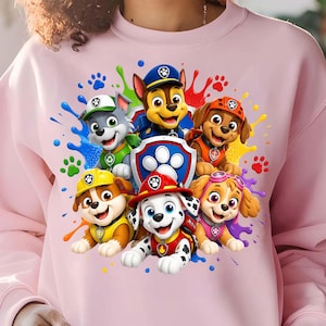 Peut inclure: Un sweat-shirt à col rond rose clair arborant une impression graphique colorée de cinq chiens de dessin animé Paw Patrol, vêtus de tenues rouges, jaunes, orange, vertes et bleues, entourant un emblème central en forme de bouclier bleu et argent.