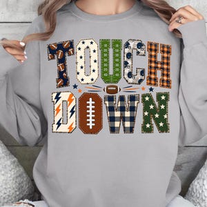May include: A light gray crewneck sweatshirt featuring the word TOUCHDOWN in large block letters. Each letter has a unique pattern including footballs, stars, a field grid, helmets, plaid, lightning bolts, and a football shape for the letter O.