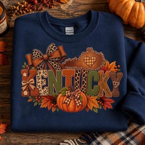 May include: A navy blue crewneck sweatshirt featuring a graphic print of the word KENTUCKY in block letters. The design includes a leopard print bow, a state outline with a plaid heart, autumn leaves, berries, and a central orange pumpkin.