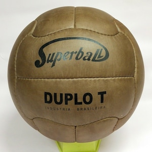 Könnte beinhalten: Ein Vintage-Fußball aus braunem Leder mit genähten Paneelen. Der Ball trägt den Text "Superball" in einer geschwungenen schwarzen Schrift und "DUPLO T" in einer fetten schwarzen Schrift. Der Text "INDUSTRIA BRASILEIRA" ist darunter aufgedruckt. Der Ball liegt auf einem grünen Ständer.
