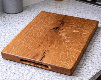 May include: A rectangular wooden cutting board with a handle cutout. The board is made of light brown wood with a visible grain pattern and a dark crack running through the center. The board is on a speckled countertop.