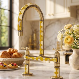Può includere: Un rubinetto da cucina a ponte con bocca alta e due manopole, rifinito in metallo dorato lucido con inserti decorativi a motivi circolari gialli e bianchi e dettagli a corda lungo i bordi della struttura del rubinetto.