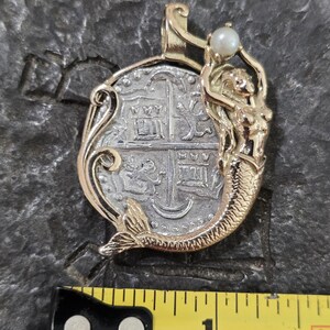 May include: A gold-toned pendant featuring a silver coin design with a mermaid figure. The mermaid holds a small pearl. The pendant is approximately 1 inch in length.