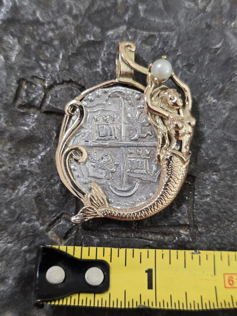 May include: A gold-toned pendant featuring a silver coin design with a mermaid figure. The mermaid holds a small pearl. The pendant is approximately 1 inch in length.