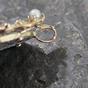 May include: Close-up of a gold-colored pendant with a small, round pearl. The pendant has an ornate design with a loop for a chain. The background is a dark, textured surface.