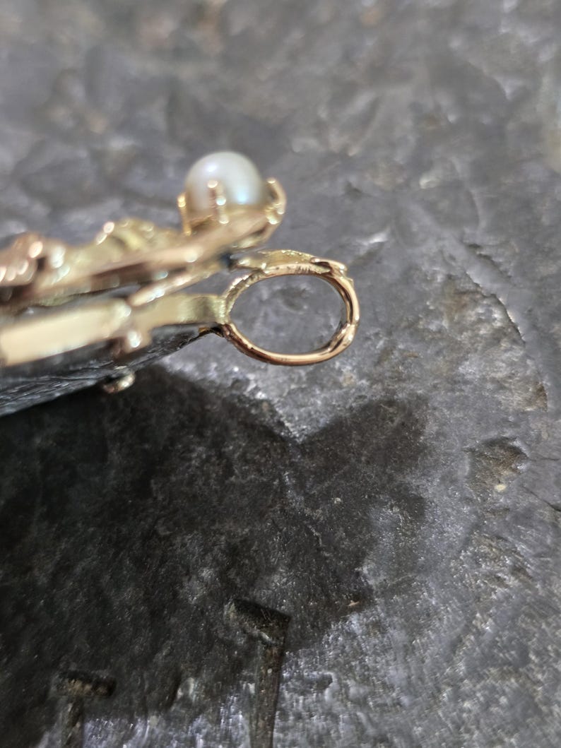 May include: Close-up of a gold-colored pendant with a small, round pearl. The pendant has an ornate design with a loop for a chain. The background is a dark, textured surface.
