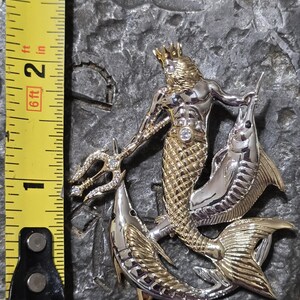 以下が含まれることがあります： 三叉槍と2匹の魚を持つ神話上の海の王様を特徴とする、ツートンカラーの金属ペンダント。ペンダントの高さは約5cmです。王様は金の王冠と腰にダイヤモンドのような宝石を身に着けています。