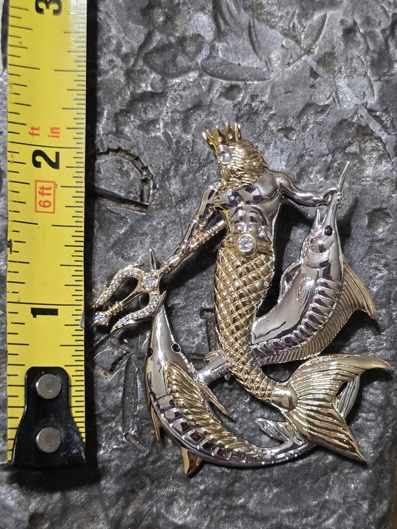 以下が含まれることがあります： 三叉槍と2匹の魚を持つ神話上の海の王様を特徴とする、ツートンカラーの金属ペンダント。ペンダントの高さは約5cmです。王様は金の王冠と腰にダイヤモンドのような宝石を身に着けています。
