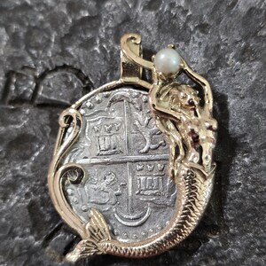 May include: A gold-toned pendant featuring a mermaid holding a pearl above a silver coin. The coin has intricate details and is framed by a gold border. The mermaid has a detailed tail and flowing hair.