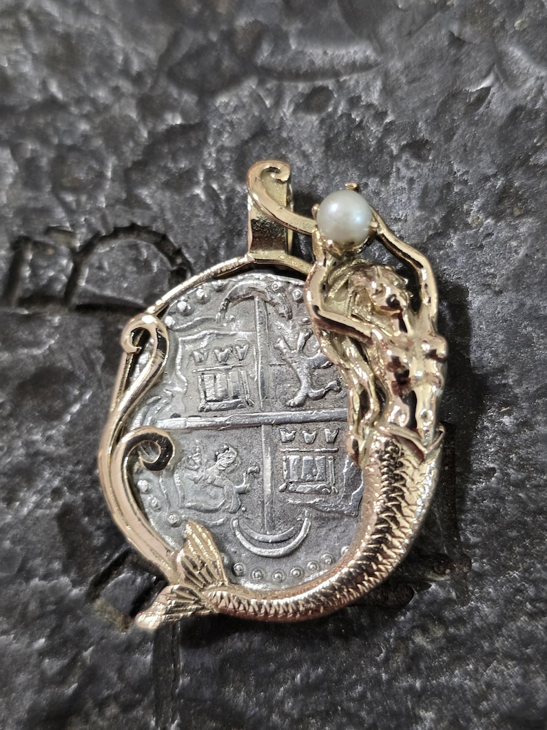 May include: A gold-toned pendant featuring a mermaid holding a pearl above a silver coin. The coin has intricate details and is framed by a gold border. The mermaid has a detailed tail and flowing hair.
