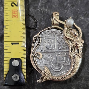 May include: A gold pendant featuring a silver coin with a detailed design. A gold mermaid holds a pearl above the coin. The pendant is approximately 1 inch in diameter, with a gold bail for a chain.
