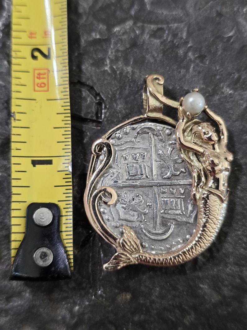 May include: A gold pendant featuring a silver coin with a detailed design. A gold mermaid holds a pearl above the coin. The pendant is approximately 1 inch in diameter, with a gold bail for a chain.