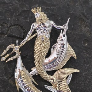 以下が含まれることがあります： 王冠、ひげ、魚の尾を持つ神話上の海の生き物を描いた、銀と金色のペンダント。 その人物は三叉の槍を持ち、2匹の魚に挟まれています。 小さな宝石がペンダントの中央にセットされています。