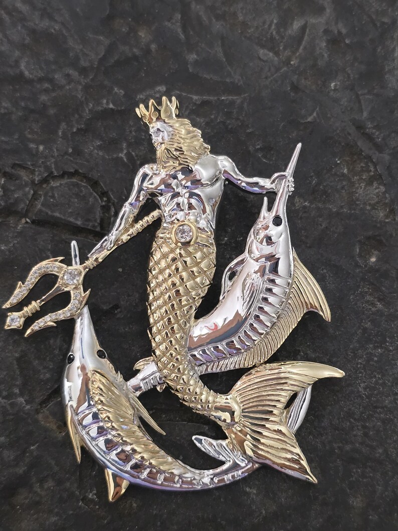 以下が含まれることがあります： 王冠、ひげ、魚の尾を持つ神話上の海の生き物を描いた、銀と金色のペンダント。 その人物は三叉の槍を持ち、2匹の魚に挟まれています。 小さな宝石がペンダントの中央にセットされています。