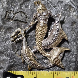以下が含まれることがあります： 王冠をかぶり、三叉の槍を持ち、2匹の魚に囲まれた、神話上の海の王を特徴とするツートーンメタルペンダント。ペンダントの長さは約5cmです。