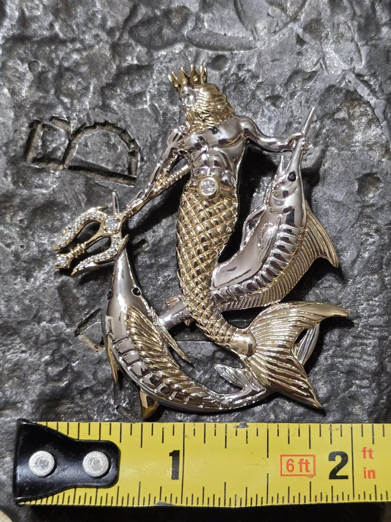 以下が含まれることがあります： 王冠をかぶり、三叉の槍を持ち、2匹の魚に囲まれた、神話上の海の王を特徴とするツートーンメタルペンダント。ペンダントの長さは約5cmです。